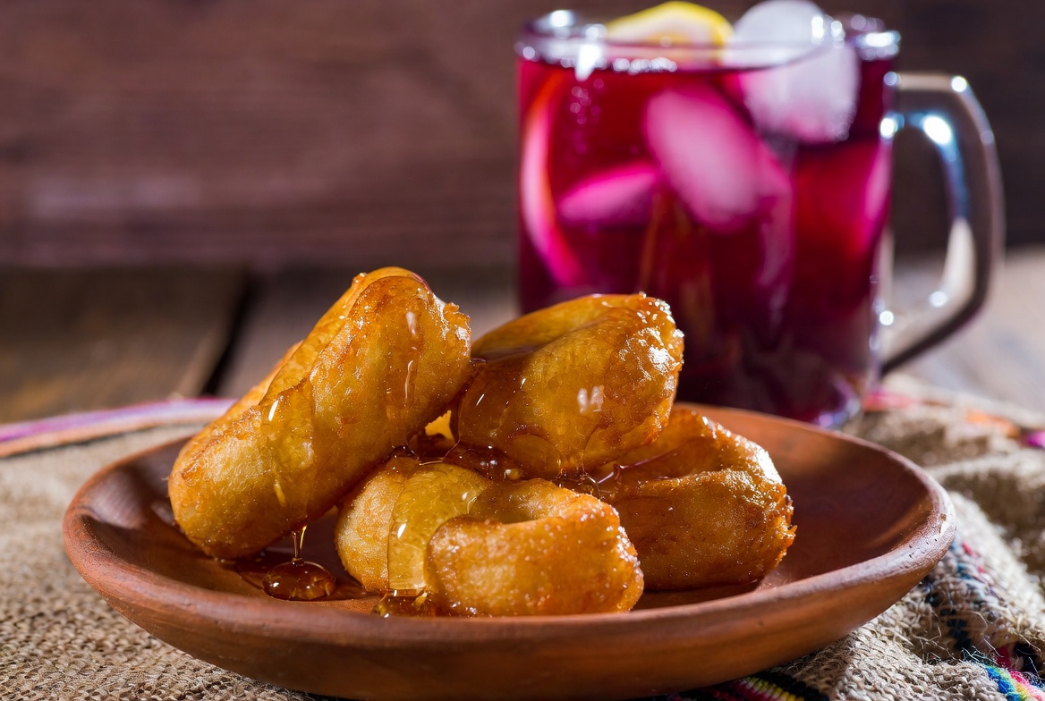 Picarones peruanos ba&ntilde;ados en miel servidos con un vaso de Chicha Morada.