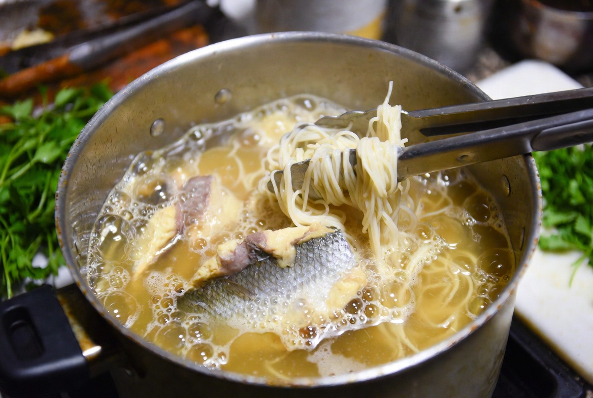 Fideos finos y trozos de pescado cociendo en el caldo dorado de la sopa