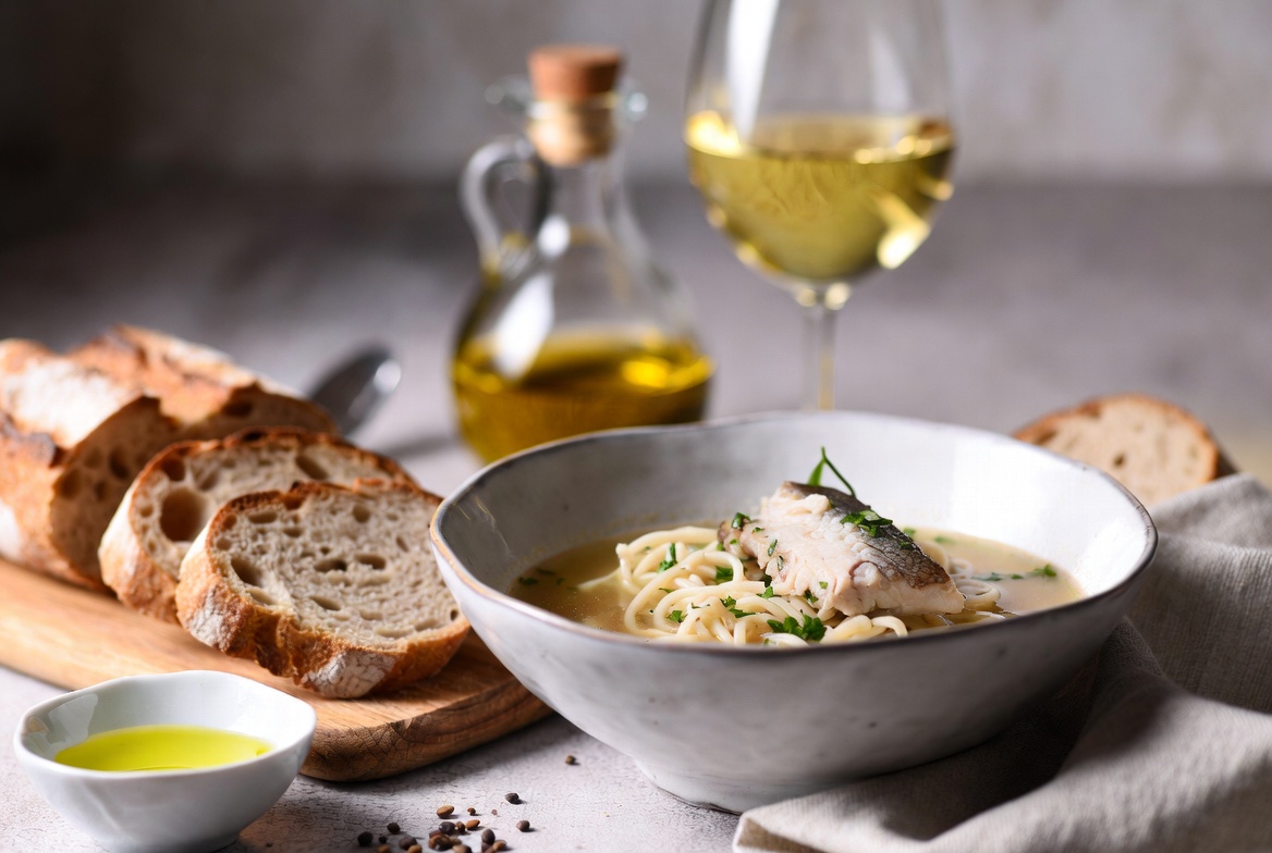 Sopa de fideos con pescado presentada en cuenco, acompa&ntilde;ada de pan r&uacute;stico y una copa de vino blanco