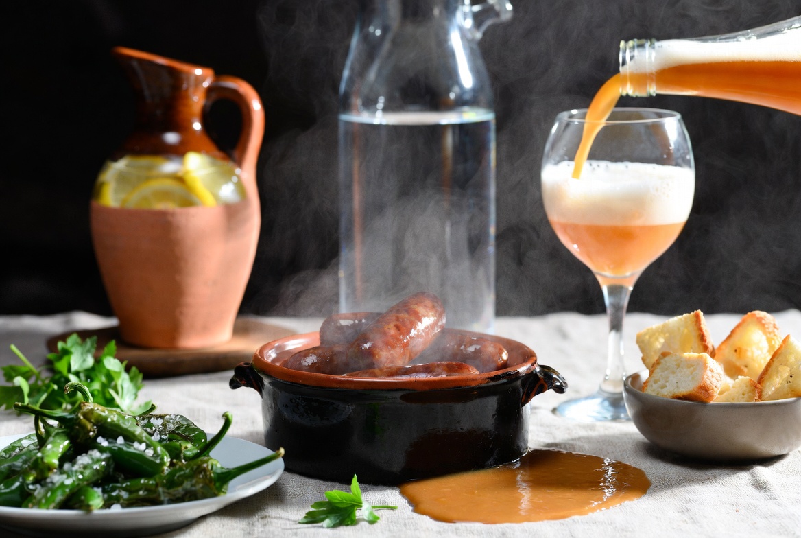 Txistorra a la Sidra en mesa con sidra natural, pimientos de Padr&oacute;n y pan para mojar